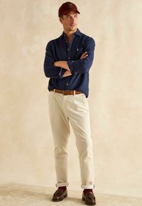 Námornícka košeľa s gombíkmi, béžové kordové nohavice, hnedé kožené topánky s tromi strapcami a bordová čiapka. Voľný strih, klasické dizajnové detaily.