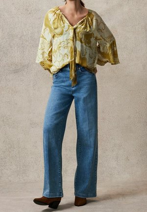 Gul og cremefarvet mønstret bluse med bindebånd foran, afslappet pasform og brede ærmer, stylet med blå, højtaljede jeans med lige ben og brune støvler.