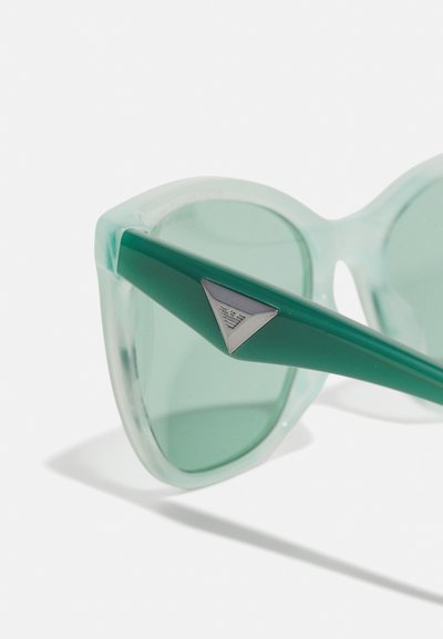 Green-tinted sunglasses with a transparent frame, cat-eye shape, and triangular silver logo accent on the side.