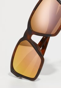 Occhiali da sole tartarugati marroni con montatura oversized e geometrica. Le lenti presentano una sfumatura che va dall'arancione al marrone. Logo discreto sulla stanghetta.