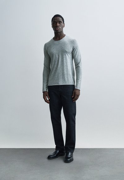 Ανοιχτό γκρι μακρυμάνικο πουκάμισο με απαλή υφή και στρογγυλή λαιμόκοψη, συνδυασμένο με σκούρο τζιν και μαύρα παπούτσια, στέκεται μπροστά σε ουδέτερο φόντο.