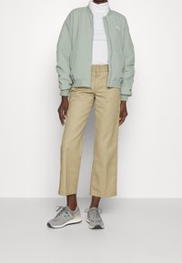 Veste bomber vert clair, col roulé blanc, pantalon taille haute beige et baskets New Balance grises avec des accents verts.
