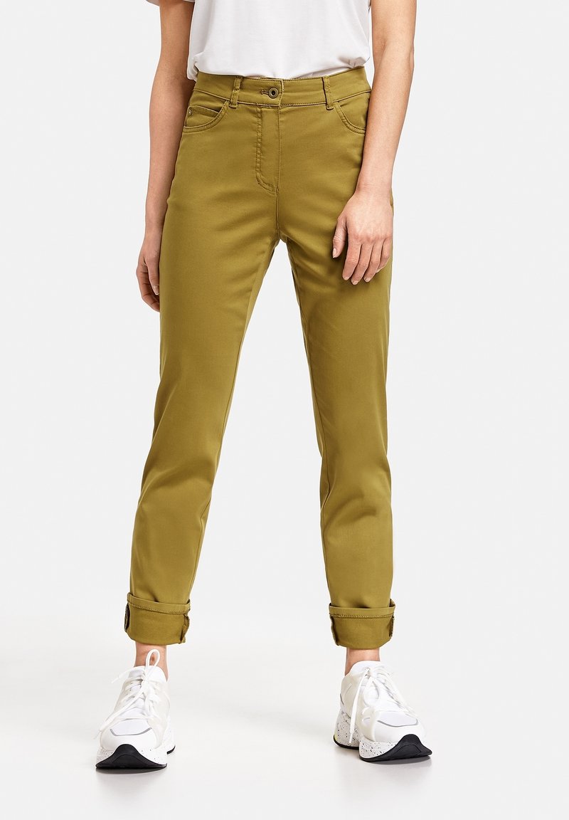 Personne portant un pantalon jaune moutarde retroussé, des baskets blanches et une chemise blanche, debout devant un fond clair et uni.