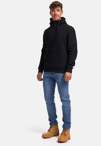 Sweatshirt à capuche noire avec un design texturé, poche avant, associée à un jean bleu et des bottes à lacets beige. Le modèle se tient sur un fond neutre.