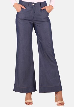 Woman wearing wide-leg dark blue denim pants with front pockets, button closure, and beige high-heeled sandals standing with hands in pockets.