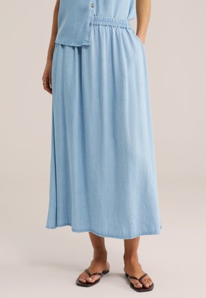 Žena oblečená v svetlomodrej midi sukni s pružným pásom a zladenom bezrukávovom topom na gombíky, doplnená čiernymi žabkami.