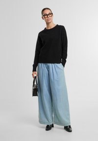 Schwarzer, strukturierter Pullover mit geripptem Saum, kombiniert mit hellblauen weit geschnittenen Jeans. Das Model hält eine kleine schwarze Handtasche und trägt elegante schwarze Schuhe.