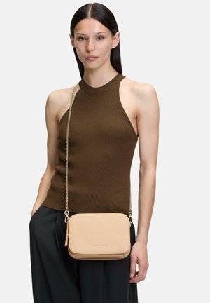 Frau mit glattem dunklem Haar, die ein ärmelloses braunes geripptes Oberteil und schwarze Hosen trägt, dabei eine kleine beige Umhängetasche mit Kettenriemen bei sich hat.