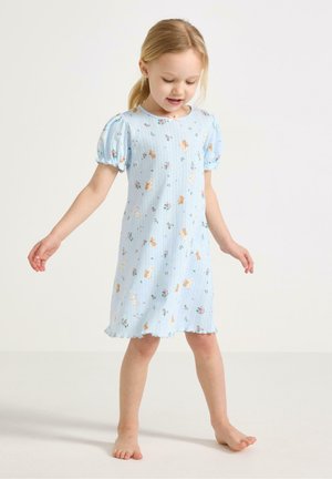 Nuori tyttö seisoo paljain jaloin, yllään vaaleansininen, lyhythihainen mekko, jossa on kukka- ja pupukuvioita valkoisella taustalla.