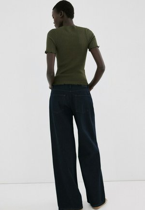 Donna in piedi con le mani sui fianchi, indossa una maglietta a maniche corte aderente verde oliva a costine e jeans larghi a gamba larga di colore blu scuro.