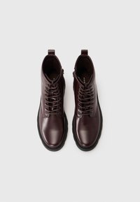 Botas de tobillo de cuero marrón con un acabado brillante, punta redonda, cordones en la parte delantera y suela negra robusta. Incluyen una pestaña trasera para facilitar el calzado.