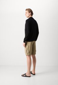 Tricou negru cu mânecă lungă, pantaloni scurți cargo crem cu buzunare și sandale negre. Material neted, croială lejeră, design minimalist, fără logo-uri vizibile.
