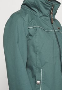 Teal waterproof jacket with a high collar, silver zipper, and contrasting stitching. Features side pockets and a smooth, textured finish.