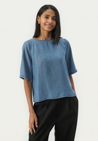 Denim modra majica s kratkimi rokavi, sproščenega kroja, okroglim izrezom in subtilno teksturo. Kombinirana z črnimi hlačami.
