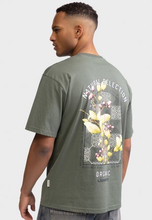 Mand iført olivengrøn T-shirt med blomstergrafik og teksten "Natural Selection" på ryggen, set bagfra og kigger mod venstre.