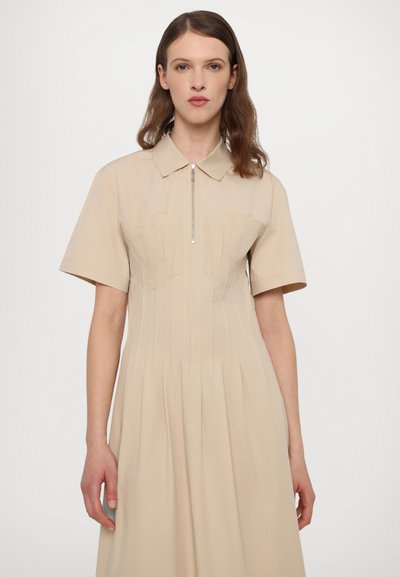 Beige mekko, jossa on lyhyet hihat, kaulus ja etuvetoketju. Sisältää laskoksia ja kaksi rintataskua rakenteellista muotoilua varten.