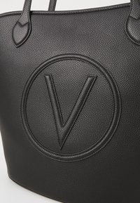 Schwarze, strukturierte Ledertasche mit einem auffälligen, geprägten "V"-Logo und kreisförmigem Rand. Verfügt über flache Griffe zum Tragen.