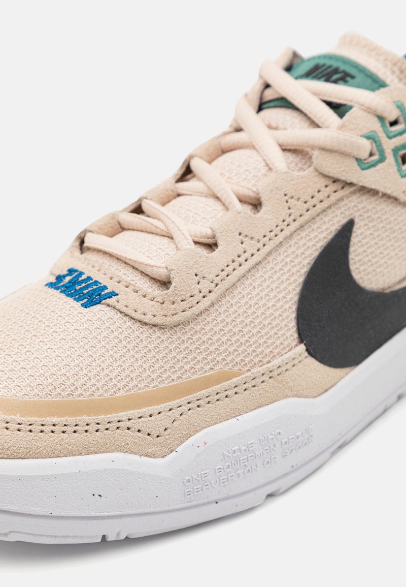 Beige Sport-Sneaker aus Mesh- und Wildledermaterialien, mit einem schwarzen Swoosh, türkisfarbenem Branding und einer strukturierten weißen Zwischensohle mit gedrucktem Text.