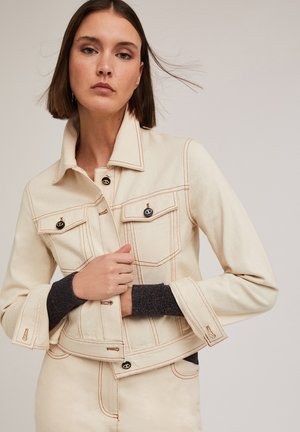Beige denim jasje met contrasterende oranje stiksels, twee borstzakken en geribbelde donkere manchetten. Voorzien van ronde zwarte knopen en een cropped fit.