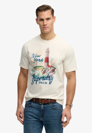 Mand iført en hvid T-shirt med print af et fyrtårn i New York, blå jeans, brun bælte og armbåndsur, stående foran en ensfarvet baggrund.