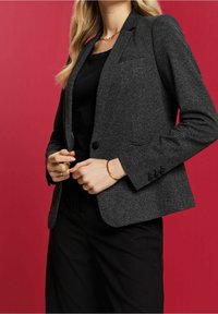 Kvinna som bär en mörkgrå strukturerad kavaj över svart topp och byxor, accessoriserad med guldsmycken, som står mot en röd bakgrund.