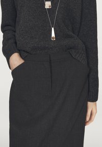 Person wearing a dark gray sweater and black skirt with hands in pockets, wearing a long silver necklace with two brown stone pendants.