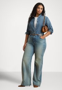 Žena v svetlom rifľovom tričku a džínsoch s hnedým opaskom a kabelkou, stojaca s jednou rukou v vrecku a neutrálnym výrazom.