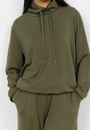 Kobieta ubrana w oliwkowo-zieloną, długorękawową bluzę z kapturem ze sznurkami oraz pasujące luźne spodnie, ręce w kieszeniach, na jednolitym tle.