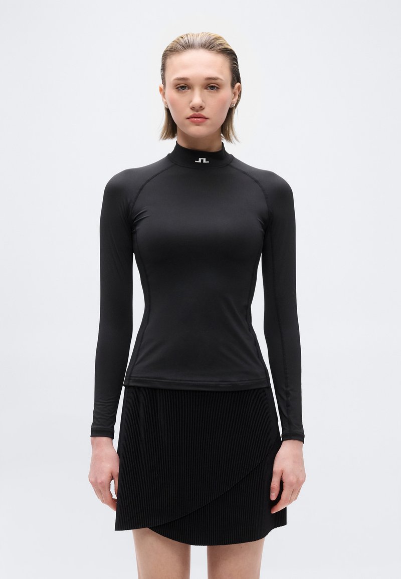 Γυναίκα με κοντά ξανθά μαλλιά που φοράει εφαρμοστό μαύρο μακρυμάνικο τοπ και μαύρη πλισέ φούστα σε απλό ανοιχτό φόντο.