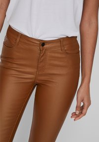 Pantalones de faux cuero marrón con un corte ajustado, un brillo sutil y detalles de costura, combinados con una camisa blanca, mostrando una mano en la cadera.