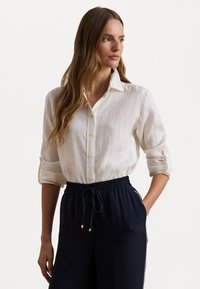 White linen button-up shirt with a relaxed fit, rolled-up sleeves, and a collar, paired with navy drawstring pants featuring gold accents.