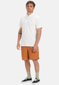 Biely polo tričko s golierom a tromi gombíkmi, kombinované s oranžovými kordurovými šortkami. Čierne tenisky s bielymi podrážkami a zelené ponožky.