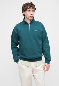 Τζελ μπλε πουλόβερ με φερμουάρ μέχρι τη μέση, με μπροστινή τσέπη και ριμπ μανσέτες. Διαθέτει μικρό λευκό λογότυπο στο στήθος. Φορεμένο πάνω από ένα ελαφρύ καρό πουκάμισο.