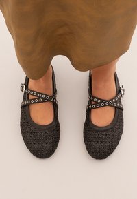 Zapatos negros texturizados con parte superior de tela trenzada, punta redonda y detalles de cuero. Presentan correas cruzadas con ojales redondos en tono plateado.