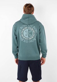 Hoodie vert avec un grand imprimé circulaire blanc comportant le nom de la marque "Oxbow" au dos. Il possède une capuche avec cordon de serrage et des poignets côtelés.
