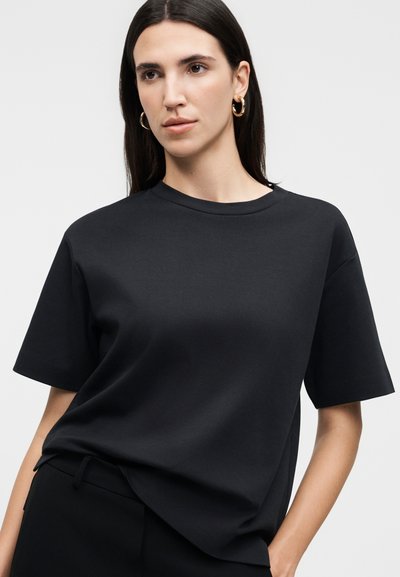 Γυναίκα με μακριά σκουρόχρωμα μαλλιά φοράει μαύρο κοντομάνικο μπλουζάκι με στρογγυλή λαιμόκοψη, χρυσούς κρίκους για σκουλαρίκια και μαύρο παντελόνι, ποζάροντας μπροστά σε φωτεινό φόντο.