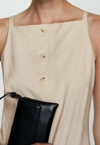 Robe beige sans manches avec un décolleté carré et trois boutons ronds ; tenant une pochette en cuir noire à la texture lisse et à la forme structurée.