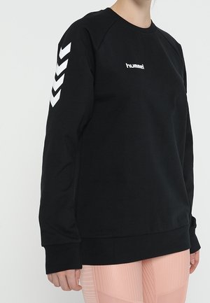 Persona que lleva una sudadera negra de hummel con franjas blancas en forma de chevrón en la manga izquierda y leggings rosa claro.