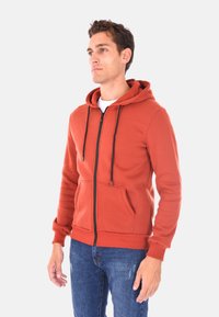 Felpa rossa con zip e cappuccio, con coulisse, tasche frontali e polsini a coste. Abbinata a jeans blu strappati. Tessuto morbido.