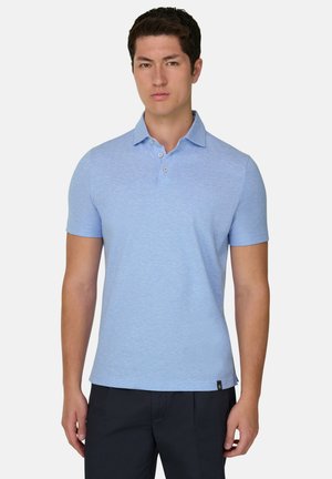 Młody mężczyzna stojący przodem, ubrany w jasnoniebieską koszulkę polo z krótkim rękawem i ciemne spodnie na jednolitym tle.