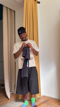 Mann trägt eine schwarze Mütze, ein Grafik-T-Shirt, weite schwarze Shorts und bunte Batik-Socken, hält ein gefaltetes schwarzes Kleidungsstück drinnen.