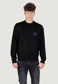 Maglione nero lavorato a maglia con maniche lunghe, scollatura rotonda e un'etichetta testurizzata con un logo sul lato sinistro del petto. Indossato con jeans scuri.