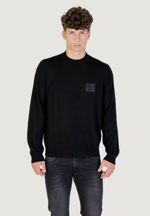 Maglione nero lavorato a maglia con maniche lunghe, scollatura rotonda e un'etichetta testurizzata con un logo sul lato sinistro del petto. Indossato con jeans scuri.