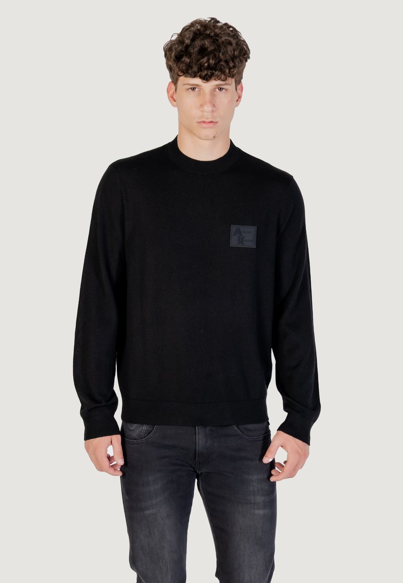 Maglione nero lavorato a maglia con maniche lunghe, scollatura rotonda e un'etichetta testurizzata con un logo sul lato sinistro del petto. Indossato con jeans scuri.