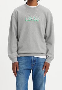 Felpa grigia con il logo "LEVI'S" in verde e bianco. Collo rotondo, maniche lunghe, polsini e orlo a coste, realizzata in materiale morbido. Indossata con jeans blu.