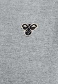 Grått tyg med en texturerad väv har en liten, broderad bi-logotyp i svart och beige, placerad på övre vänstra sidan.