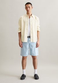Lichtgele overhem shirt, witte tanktop, lichtblauwe denimshorts, zwarte riem en zwarte instappers met witte sokken. Eenvoudige, casual outfit.
