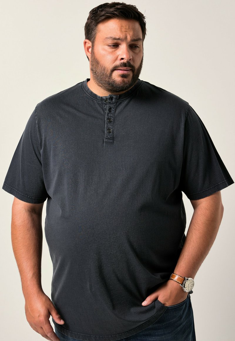 Muž s vousy, oblečený v černém tričku s krátkým rukávem a hnědých hodinkách, dívající se dolů s jednou rukou v kapse na jednobarevném pozadí.