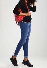 Roter Leder-Rucksack, schwarzes Langarmoberteil, blaue enge Jeans mit umgeschlagenem Saum und schwarze Sportschuhe. Lockige Haare sichtbar.