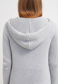 Světlounce šedá pletená mikina s kapucí a texturovaným vzorem, se stahovací šňůrkou na kapuci a volným střihem, pohled zezadu.